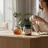 Asia Trade blackcurrant leaf herbal tea jar on a wooden table with a woman drinking tea in a bright room