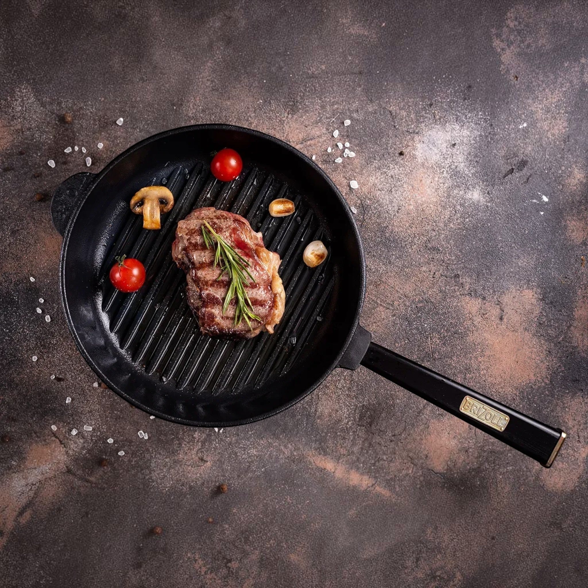 Asia Trade BRIZZOL cast iron grill pan with wooden handle, featuring a seared steak, mushrooms, and cherry tomatoes.
