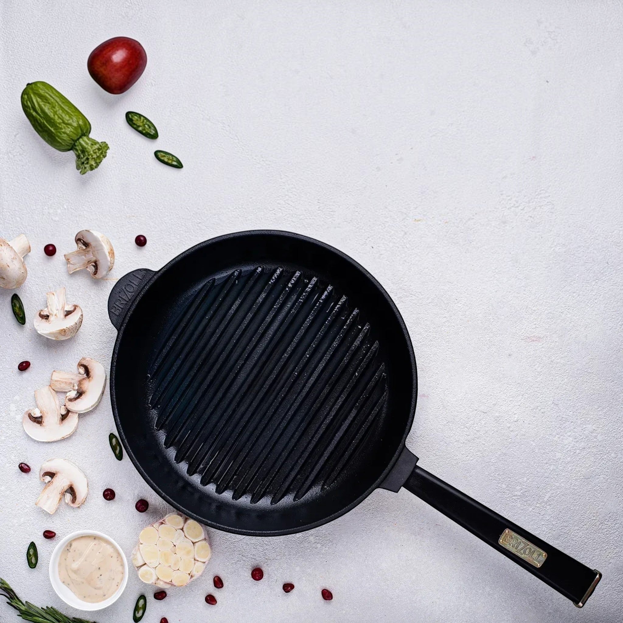 Asia Trade BRIZZOL 26cm cast iron grill pan with wooden handle, surrounded by fresh mushrooms, garlic, and peppers.