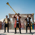 Asia Trade performers in traditional Uzbek attire playing a long golden karnay trumpet and drum at an outdoor event.