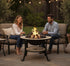 Asia Trade round mosaic fire pit table with safety mesh, used by a couple roasting marshmallows in a garden setting.